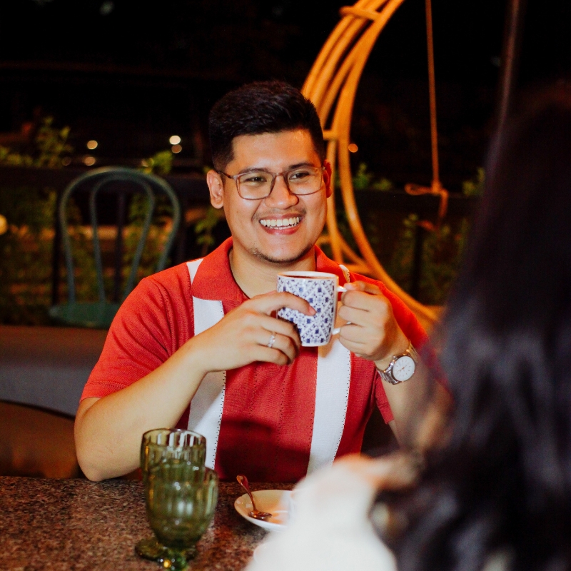 A man in a red and white shirt smiles while holding a patterned coffee mug at an outdoor gathering. He is seated at a table with a green glass and plate in front of him, enjoying the moment in a cozy, dimly lit environment.