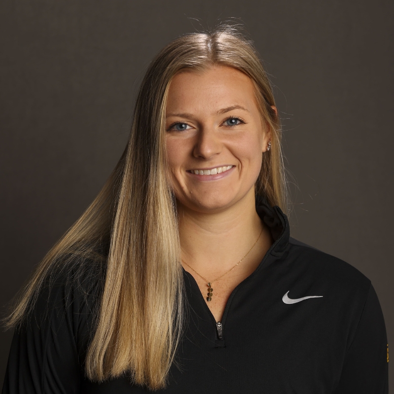 A young woman with long blonde hair, wearing a black Nike pullover and a delicate necklace, smiles softly in front of a neutral background.