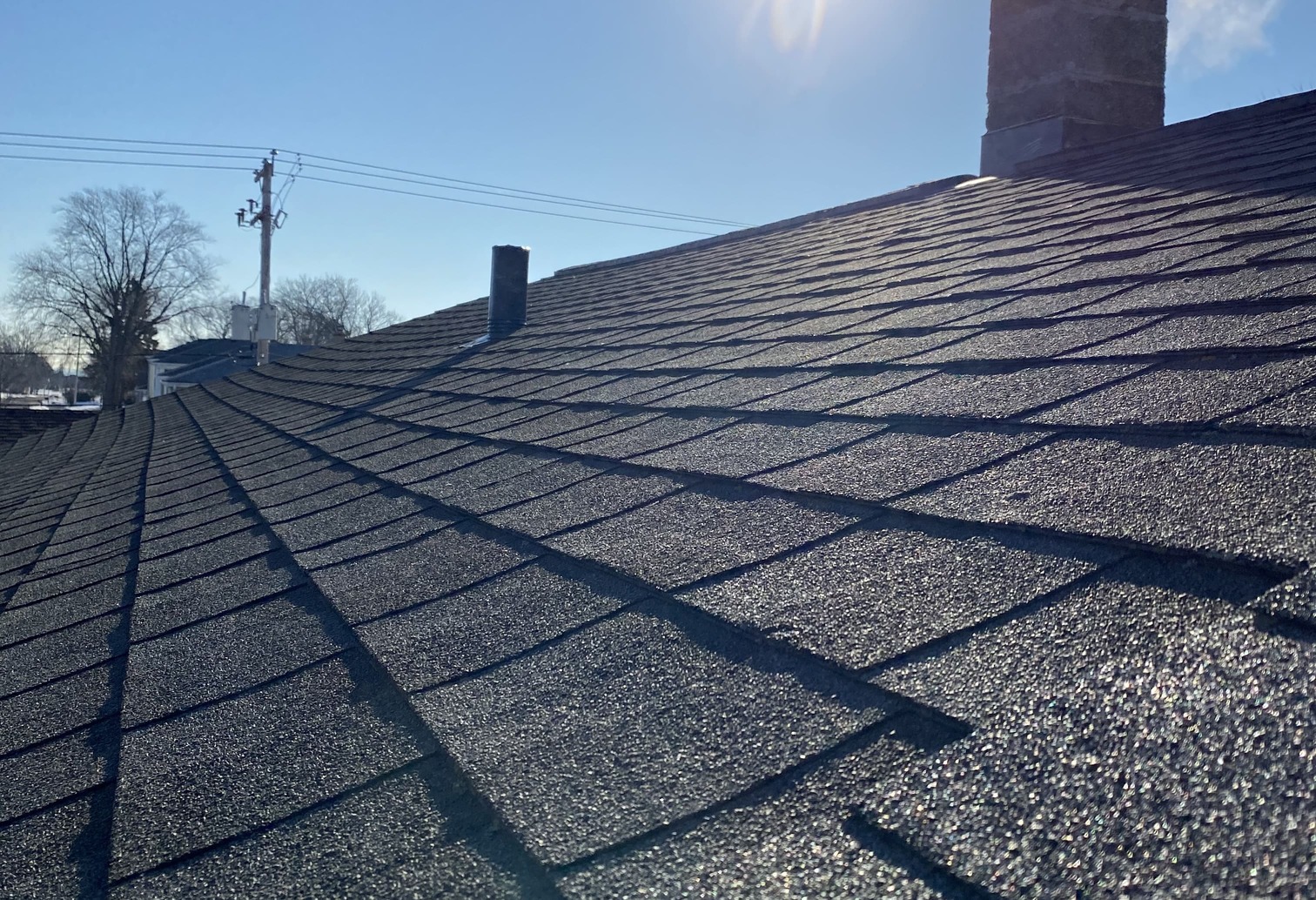 A repaired asphalt shingle roof under a clear blue sky, with sunlight highlighting the even surface. A chimney and vent are visible on the rooftop.