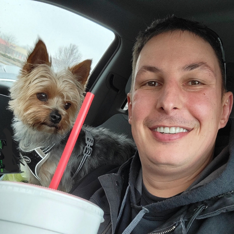 A man smiles while sitting in a car with a small Yorkshire Terrier on his shoulder. He is holding a large cup with a red straw, and the dog is looking out of the window. Both appear content and relaxed.