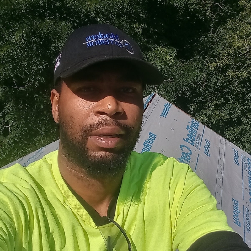 A man wearing a bright yellow work shirt and a black "Modern Exterior" hat takes a selfie while working on a roofing project. Behind him is part of the roof covered in protective material, and trees are visible in the background. Modern Exterior